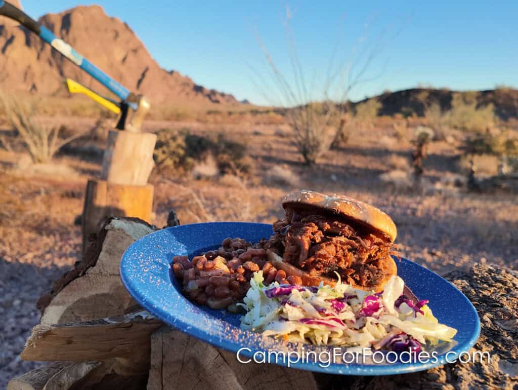 Shredded BBQ Beef Sandwiches by CampingForFoodies features a blue camping plate set on a campfire log. The plate is filled with a BBQ beef sandwich, baked beans and coleslaw tossed in a creamy dressing.