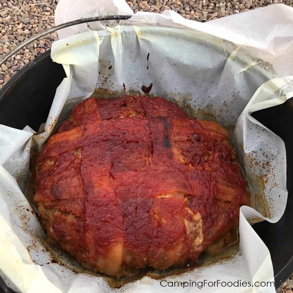 Dutch Oven Meatloaf by CampingForFoodies features a bacon and glazed-topped meat loaf nestled in a Dutch oven parchment paper liner that is set inside a cast iron camp Dutch oven ready to be sliced and served at the campsite.